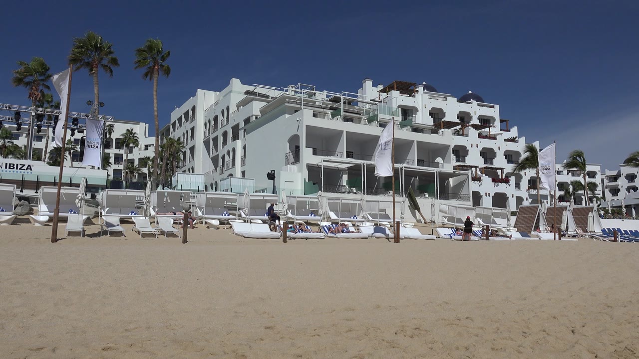 Cabo San Lucas beach view with the central white building prominent, lounge chairs arranged along the sand, palms on the left, flags in front, and tiny people in the distance.