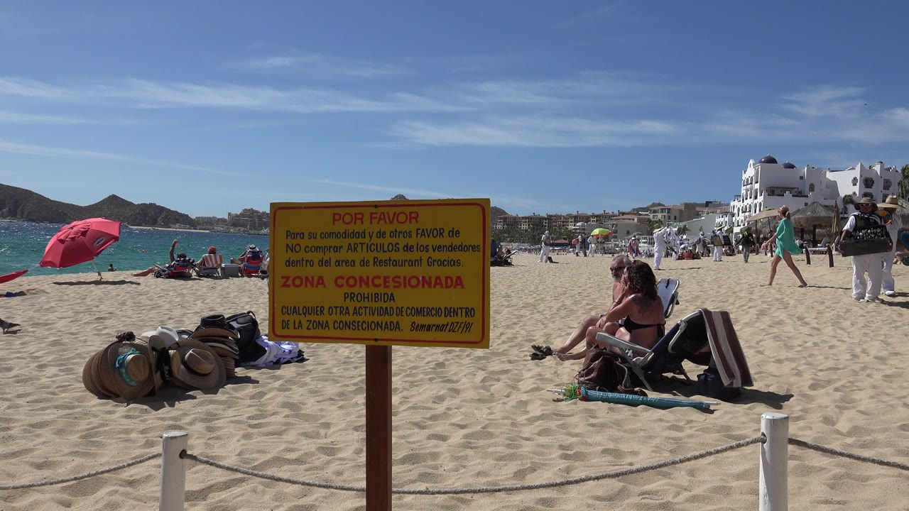 Cabo San Lucas beach frame with a yellow Spanish concession sign in the foreground, straw hats on sand, seated visitors, a pink umbrella, blue water, white buildings, and low hills behind them.