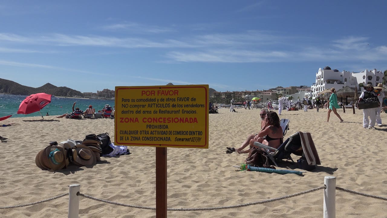 Cabo San Lucas beach view with the yellow sign centered, straw hats and towels low on the sand, adults reclining at right, people walking near the water, and white buildings below the hills.