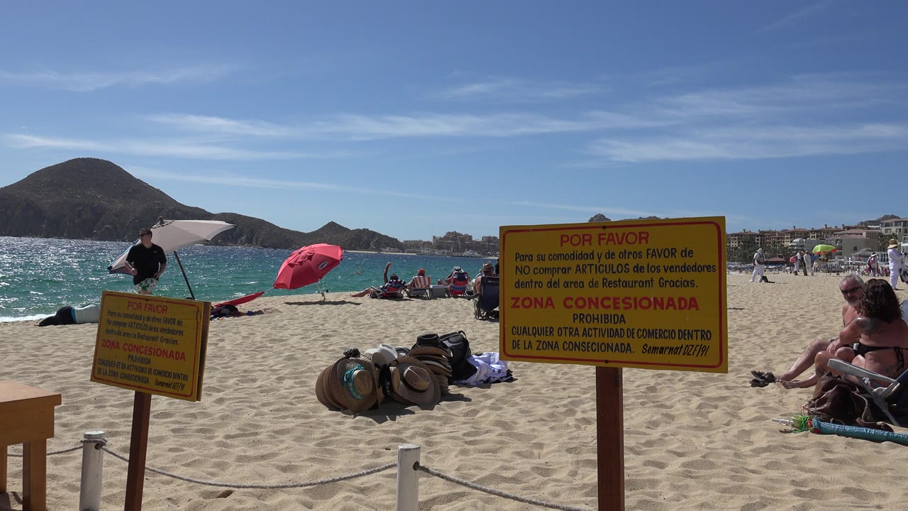 Beach sign, umbrellas, tourists, sand, water, and mountains.