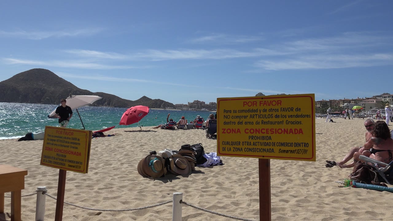 A man in dark clothing walks along the Cabo San Lucas sand near a yellow sign while umbrellas, beach bags, seated people, water, and mountains fill the background.