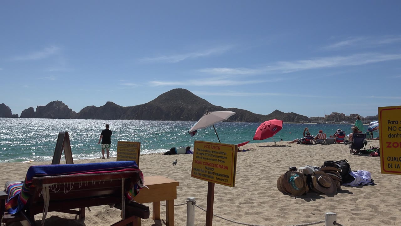 Opening frame of a Cabo San Lucas beach with striped foreground furniture, yellow signs, bright ocean glare, rocky offshore landforms, umbrellas, and distant beachgoers.