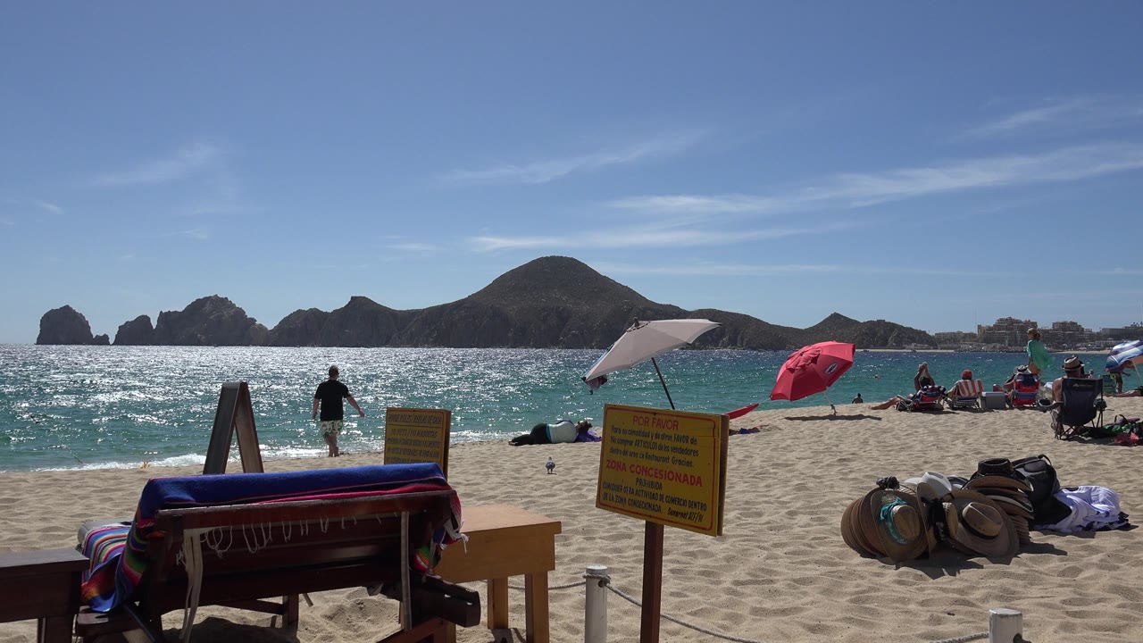 Beach foreground with striped furniture and signs facing sunlit water and rocky islands.