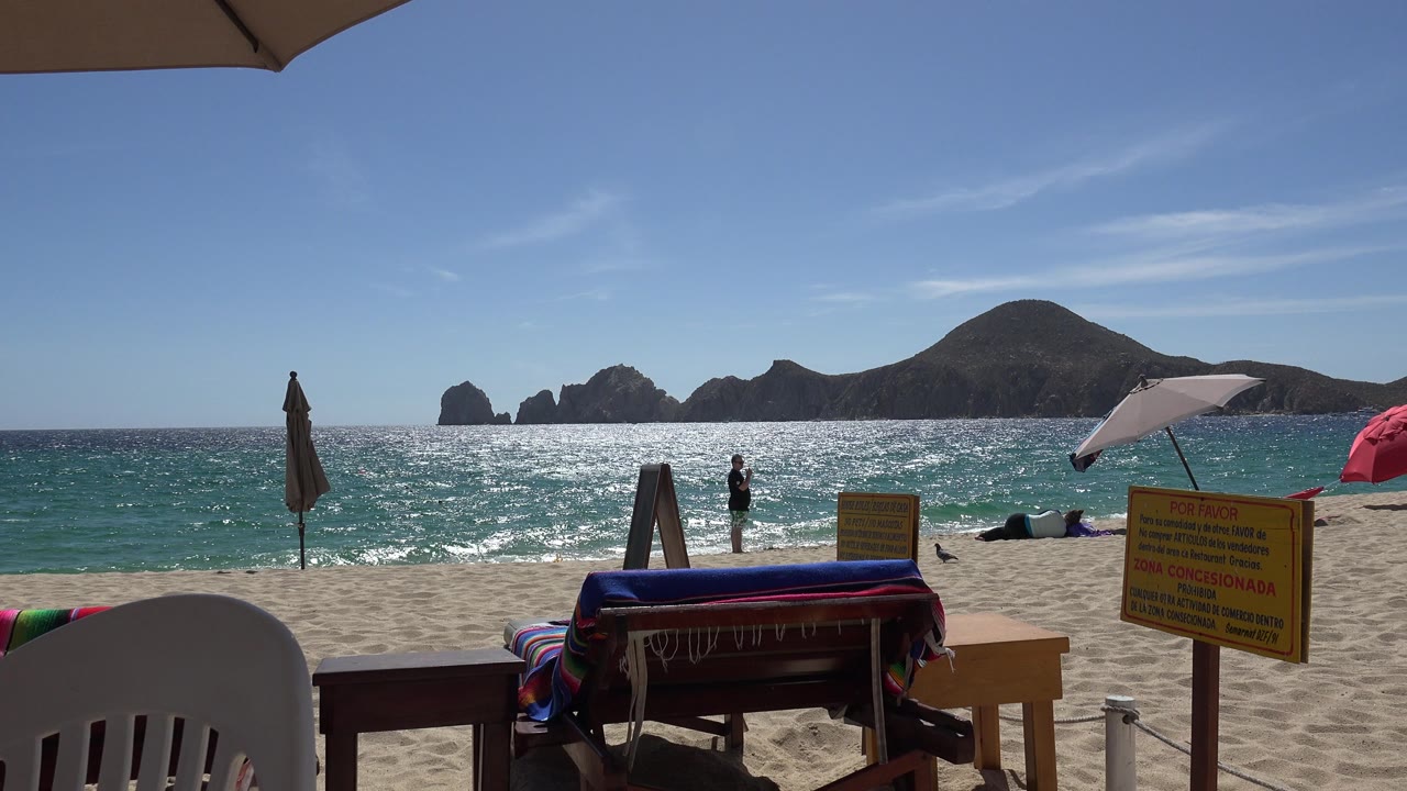 Sunny beach view with ocean, umbrellas, chair, signs, and distant rocky landforms.