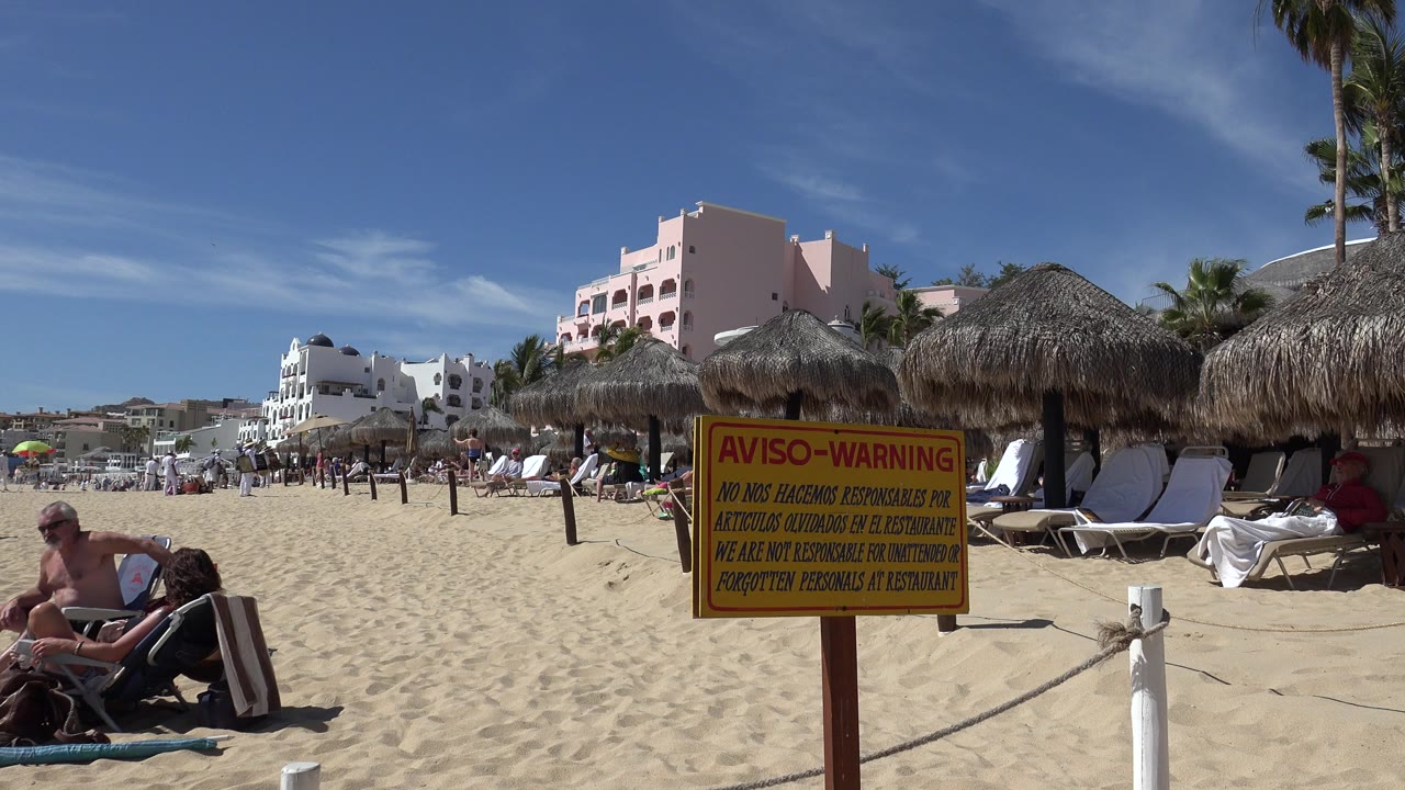Busy sandy beach with warning sign, palapas, lounge chairs, and resort buildings.