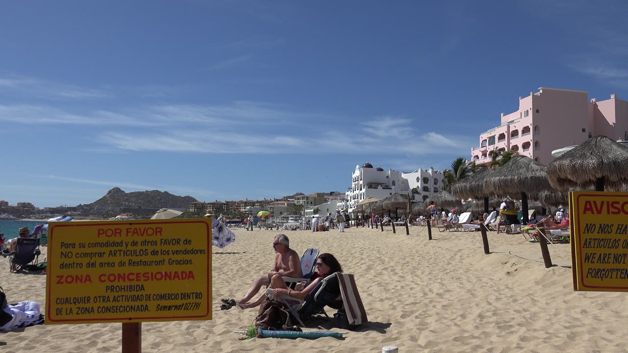 Sunny beach with seated visitors, warning signs, ocean, and resort buildings.