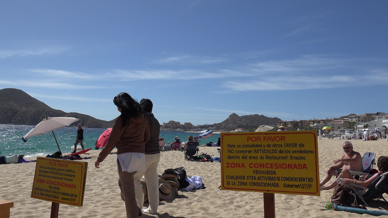 Cabo San Lucas beach scene with two walkers in the foreground, yellow warning signs on the sand, turquoise water, seated visitors, umbrellas, and low mountains behind them.