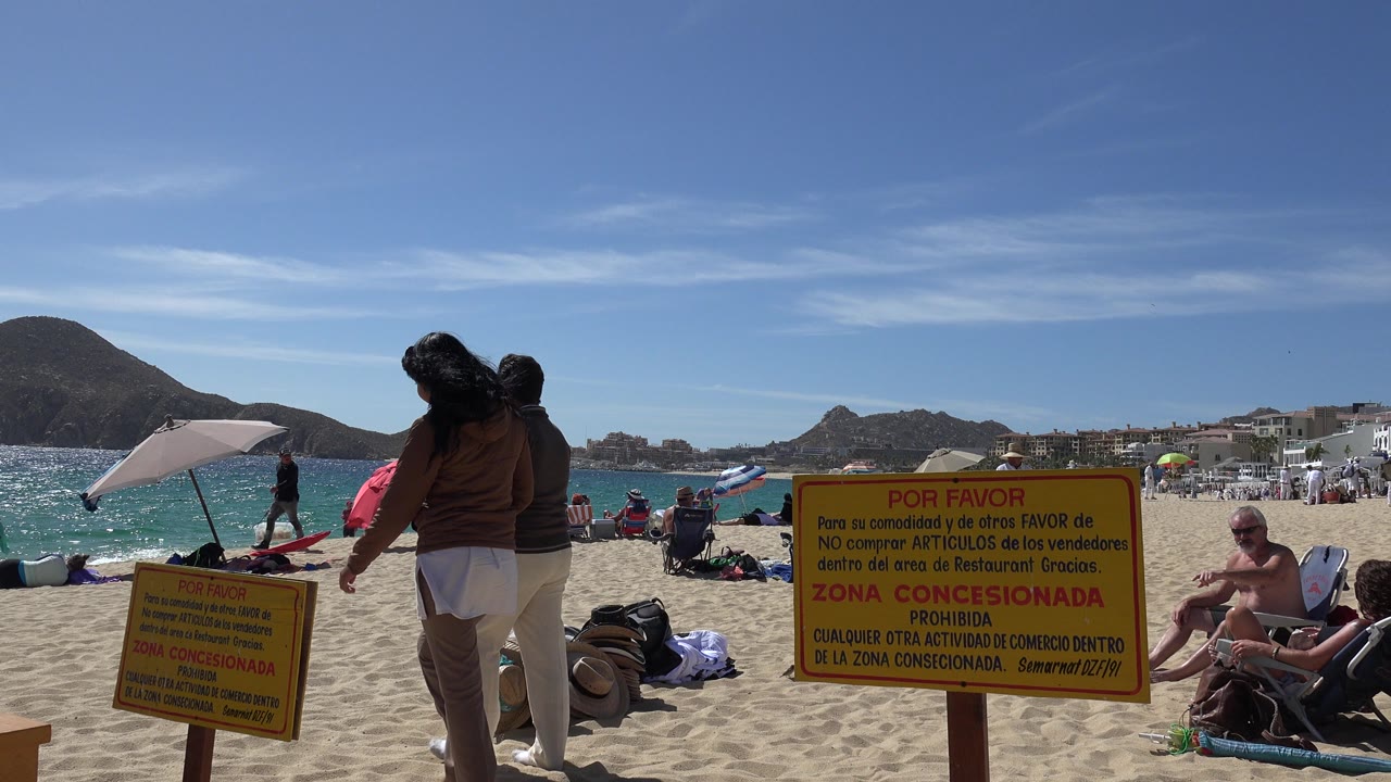 Cabo San Lucas beach frame with foreground walkers, beach bags and towels on the sand, a yellow concession warning sign, turquoise surf, umbrellas, seated visitors, and mountains in the distance.
