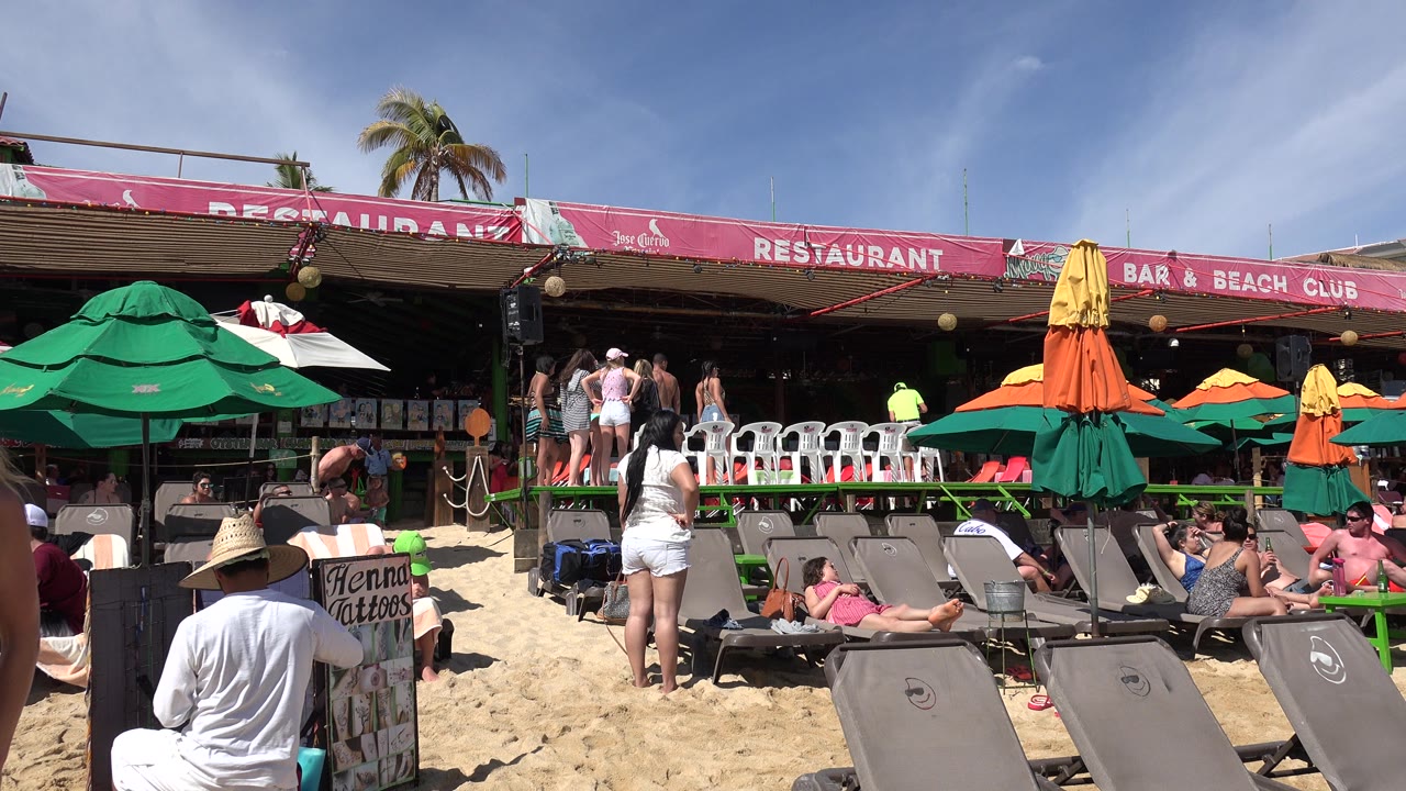 Cabo San Lucas beach club view with a seated person in a straw hat in the foreground, sunbathers on loungers, and people standing on the restaurant deck.
