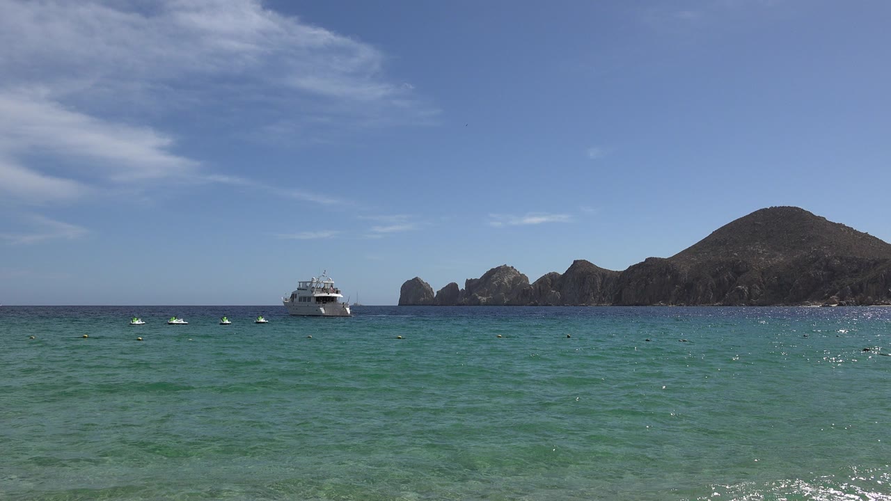 Opening frame of Cabo San Lucas, Mexico, with turquoise water in the foreground, a small white boat left of center, rocky outcrops and a dark hill in the distance, and no visible people.