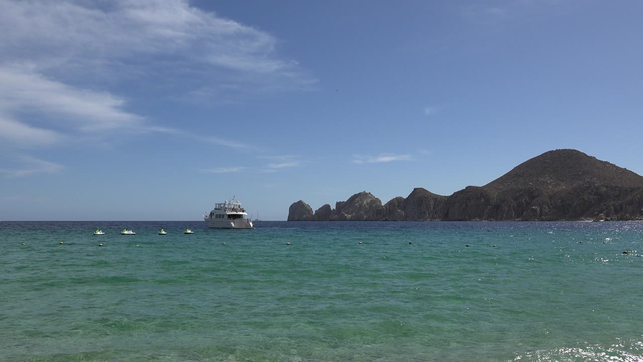 Cabo San Lucas ocean frame with the white boat slightly left of center, small floats spread across the water, rocky formations behind it, a low hill to the right, and no visible people.