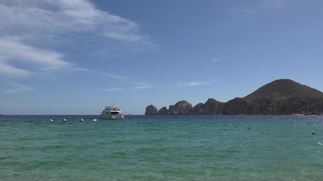 Cabo San Lucas green sea fills the foreground with a white boat left of center, rocky hills in the distance, thin clouds overhead, and no visible people in the foreground.