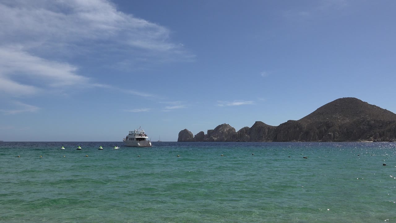 Cabo San Lucas shoreline view with green water in front, small distant water users or markers near the horizon, a white boat offshore, and rocky hills beneath a bright blue sky.