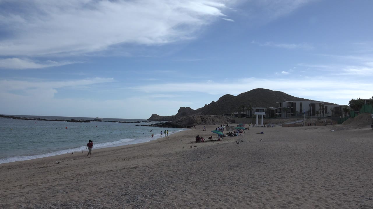 Wide sandy beach with small waves, scattered people, rocky hills, and blue sky.