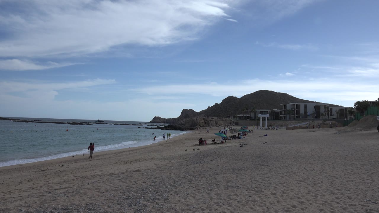 Cabo San Lucas beach view at 0:01, with a person standing near the waterline, groups farther down the sand, gentle surf, rocky hills, and beachfront structures under streaked clouds.