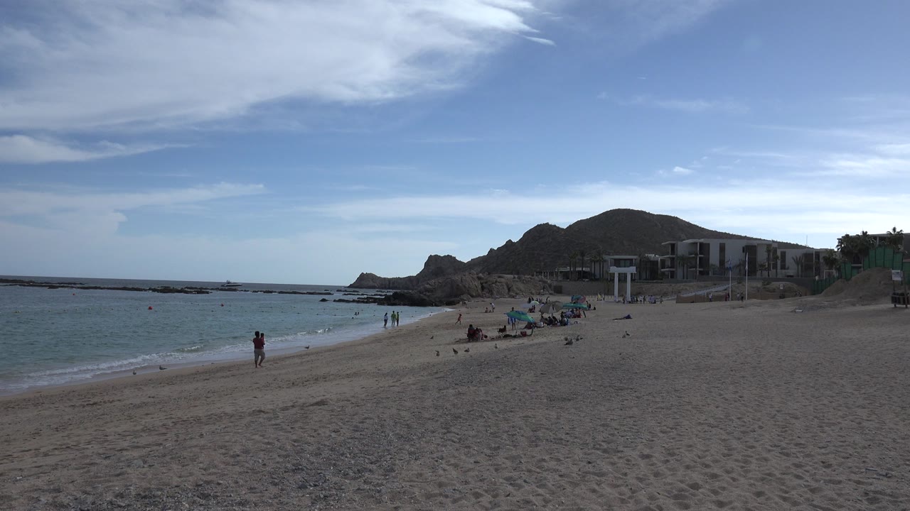 Wide Cabo San Lucas beach view at the start of the clip, with a sandy foreground, small surf at left, distant people near the waterline, rocky hill, and low buildings beyond the beach.