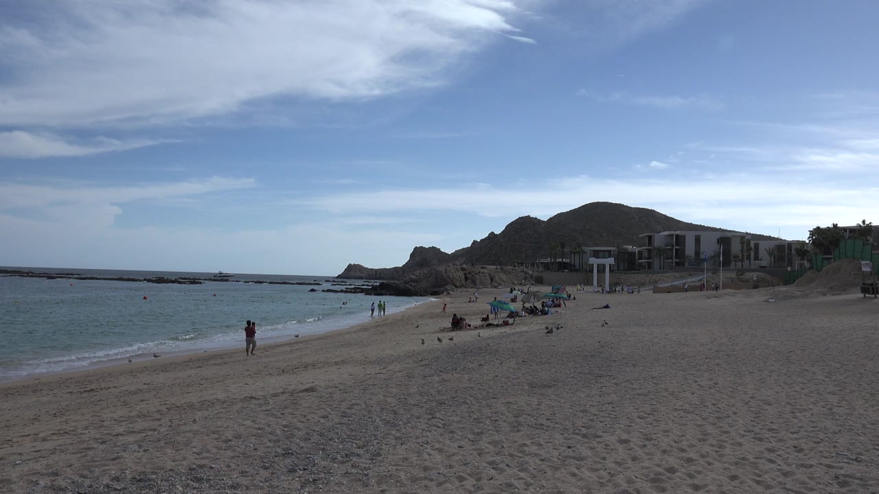 Cabo San Lucas shoreline one second in, showing a couple walking near the surf, a few other beachgoers, umbrellas on the sand, blue-green water, and rocky hills behind beachside structures.