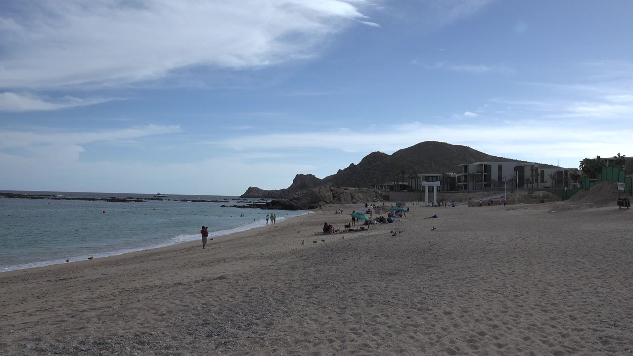 Wide beach with water, sand, distant people, buildings, and rocky hills.