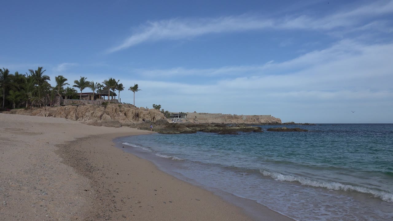 Cabo San Lucas beach at the start of the clip, with pale sand in the foreground, blue-green water at right, rocky coast and palm trees beyond, and no visible people.