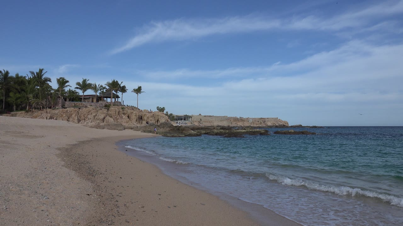 One second in, a small wave reaches farther onto the Cabo San Lucas sand while the rocky point, palms, and open blue sky remain in view with no visible people.