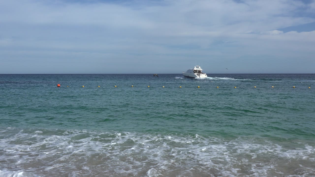 A white motorboat sits left of center on the Cabo San Lucas horizon beyond blue-green water, yellow buoys, foamy foreground surf, and no visible people.