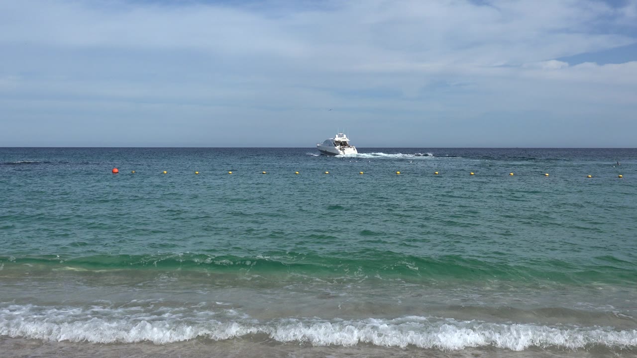 Cabo San Lucas beach view at the start of the clip, with white surf in the foreground, a buoy line across the water, a distant white speedboat near the horizon, and no visible people.