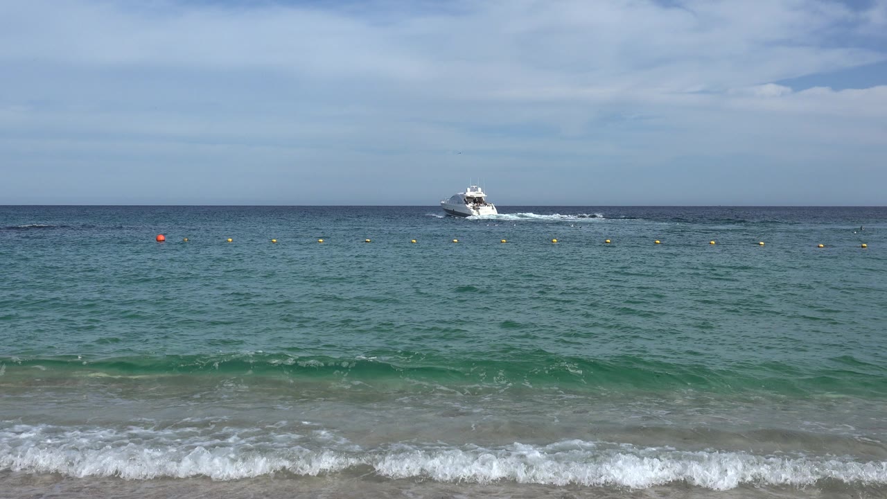 Cabo San Lucas ocean frame with the white speedboat slightly farther across the horizon, small buoys spanning the midwater, soft clouds overhead, and no visible people.