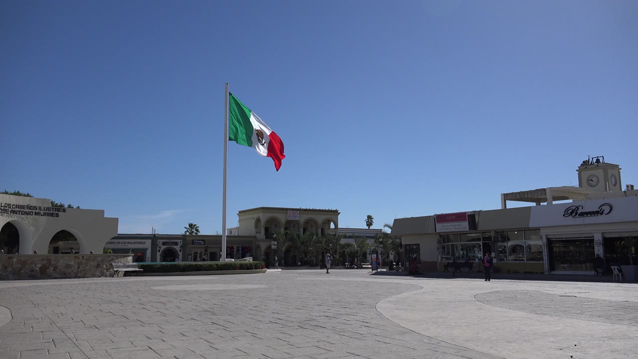 Wide view in Los Cabos, Mexico, with a Mexican flag on a tall pole at left, storefronts across the square, and small pedestrians visible near the buildings.