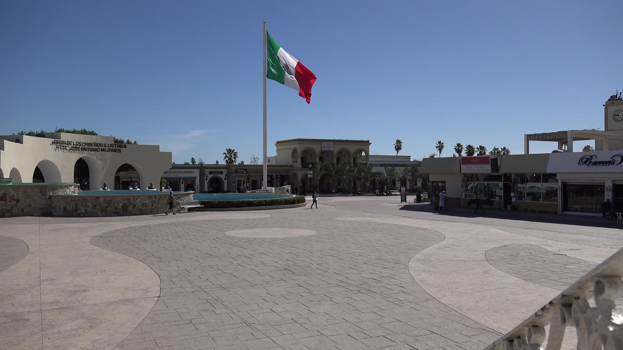 Mexican flag above a quiet paved square with arches and shops
