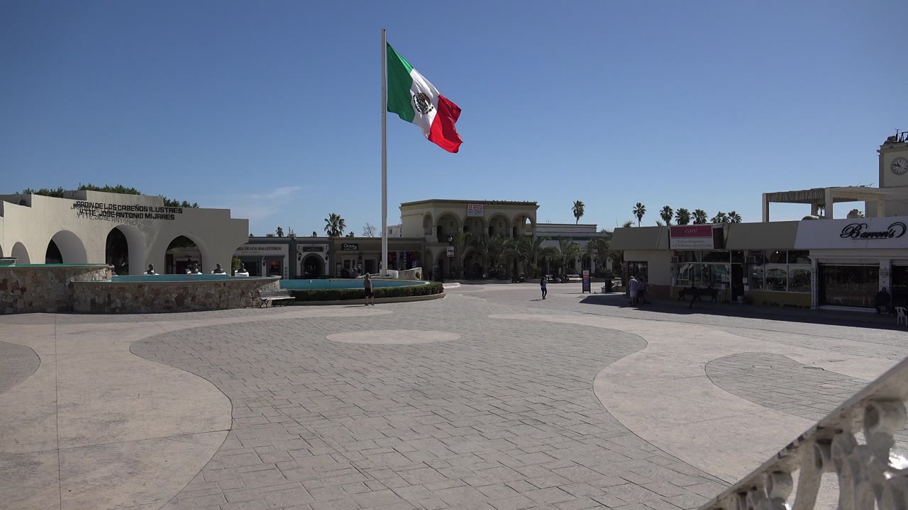 A Mexican flag flies on a tall pole above a sunny paved plaza in Los Cabos, Mexico, with arched white buildings, a low fountain or planter, storefront signage, and distant people.
