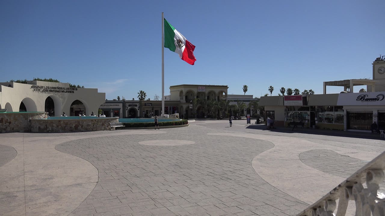 The Mexican flag remains high over the Los Cabos plaza, with the white arched facade at left, a shaded storefront at right, palm trees along the roofline, and a few distant pedestrians.