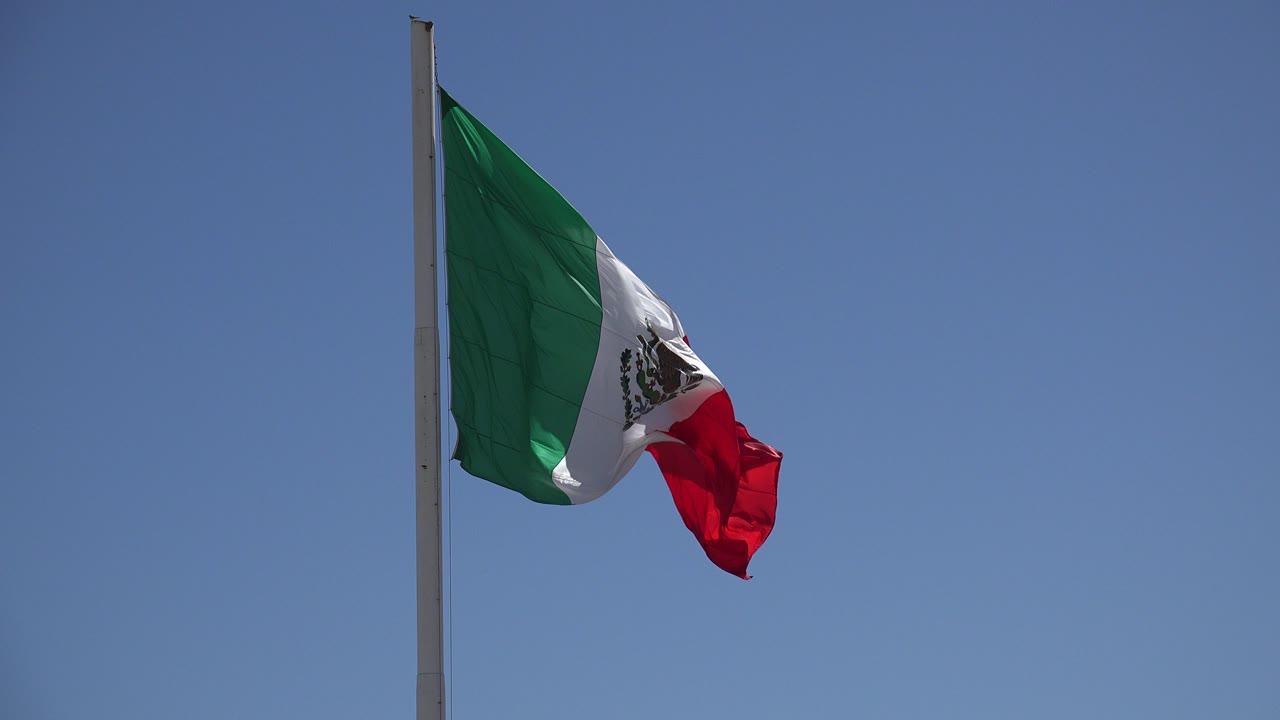 A Mexican flag in Los Cabos, Mexico folds tightly in the wind near the top of a tall pole, with blue sky behind it and no visible people.