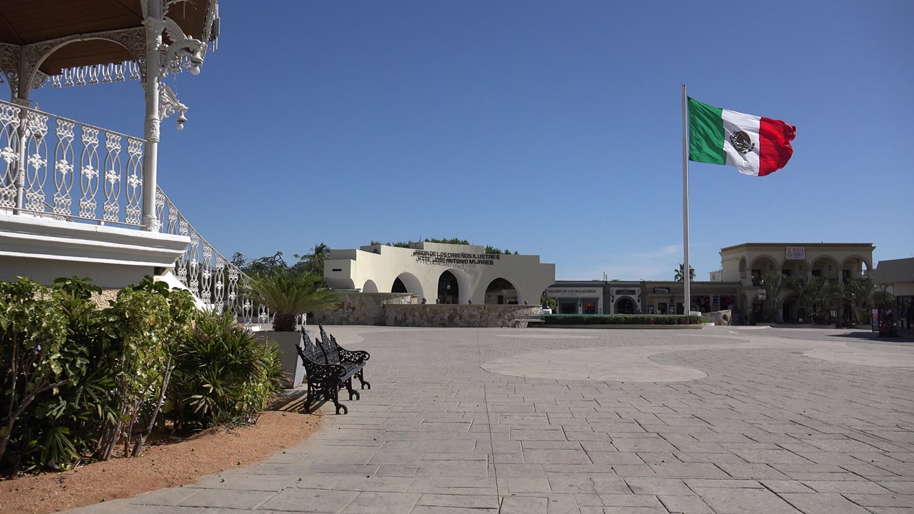 Opening view in Los Cabos, Mexico, with a Mexican flag waving above a paved plaza, white balcony railing at left, arched buildings in back, and no visible people.
