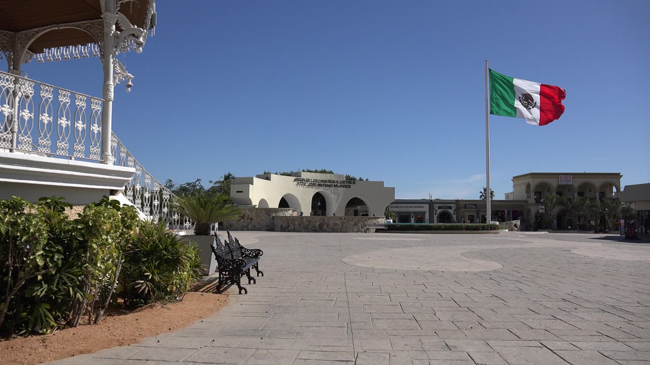 One second in, the Mexican flag is extended by the wind over the Los Cabos plaza, with the foreground balcony and bench still visible and no visible people.