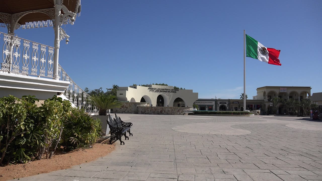 Opening frame of a Los Cabos plaza with foreground plants, a black bench, white balcony railings, arched buildings, a Mexican flag, and no visible people.