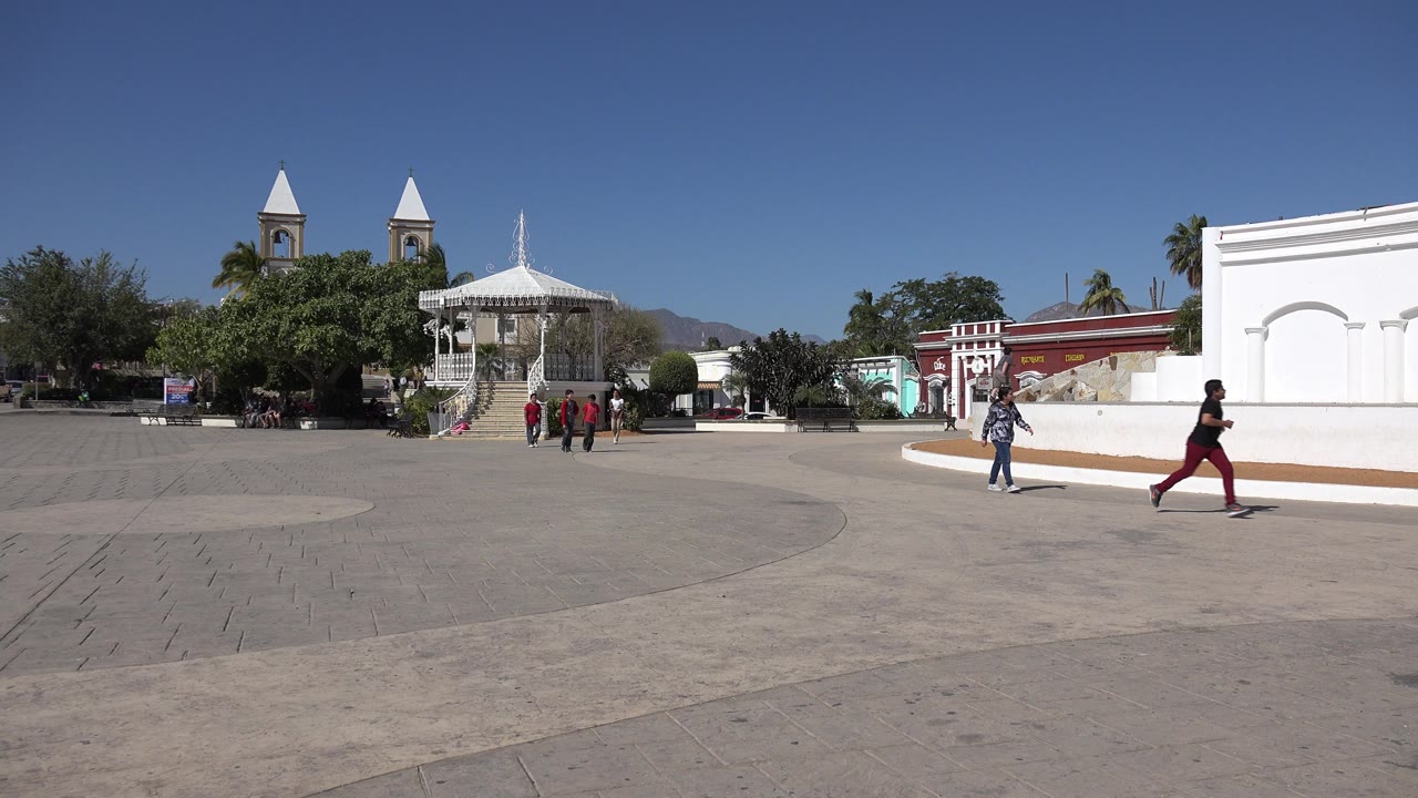 A group walks across the midground of a Los Cabos plaza while one person in dark clothing and red pants crosses near the white wall on the right.