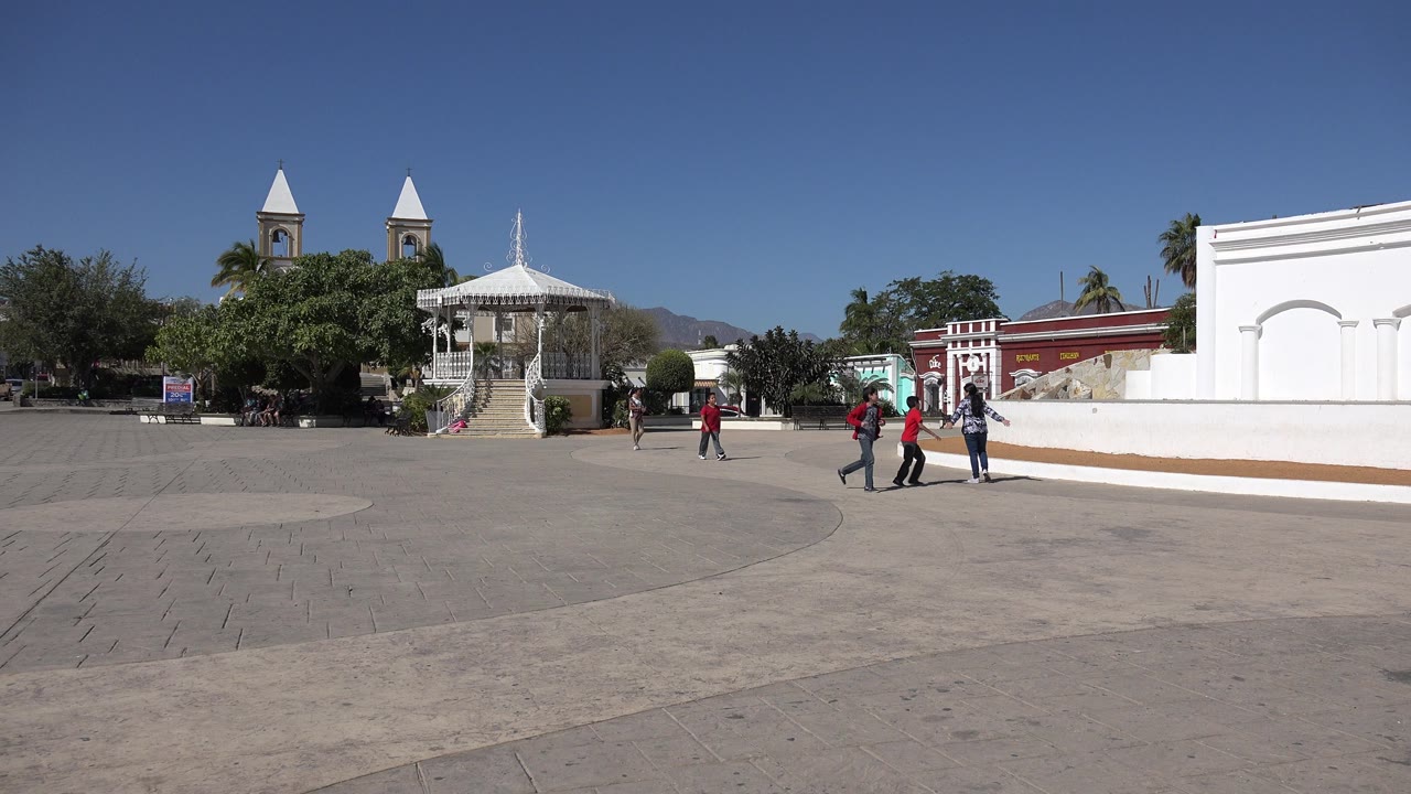 Wide view of a concrete plaza in Los Cabos, Mexico with trees, a white gazebo-like structure, twin tower-like structures, a white wall, blue sky, and several pedestrians crossing the open space.