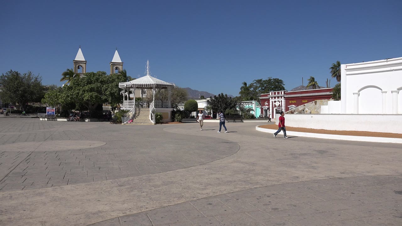 Wide public square with gazebo, trees, church-like towers, and pedestrians