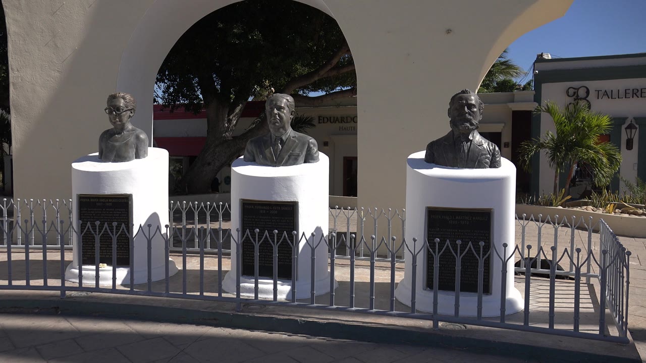 The camera view holds on the busts and plaque-fronted pedestals in Los Cabos, with a large shaded tree behind the arches and no visible people.