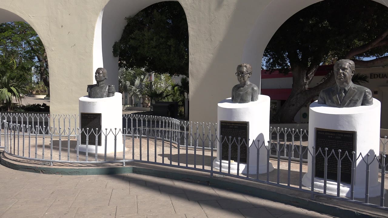 Bronze busts on white pedestals stand behind a low metal fence in Los Cabos, with cream arches, trees, palms, and hard sunlight across the pavement.