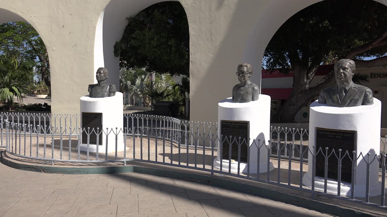 Three bronze busts in Los Cabos are visible behind a curved fence, with a central bust on a white pedestal, shaded trees, and bright sun on the stucco arcade.