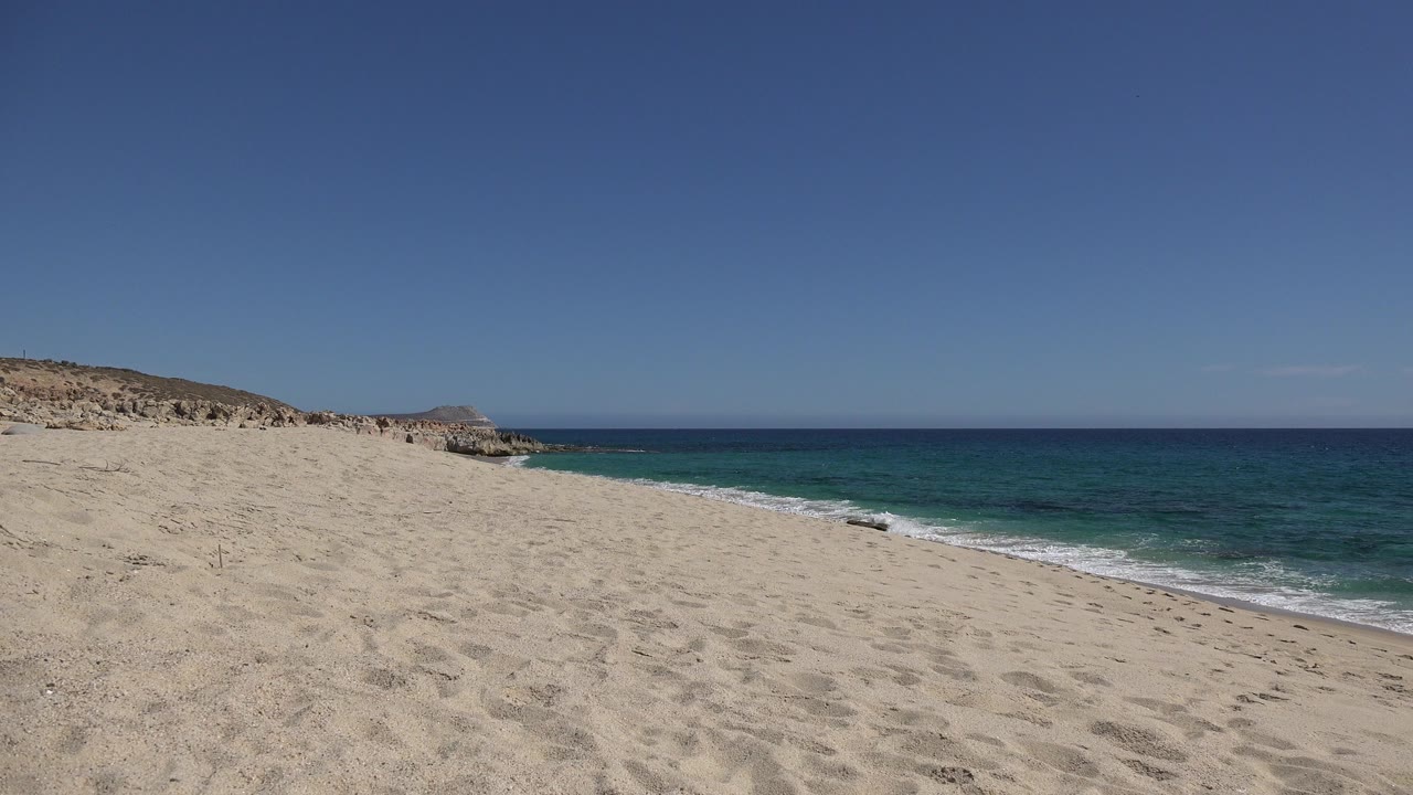 Opening frame of a Los Cabos, Mexico beach with footprint-marked sand in front, blue water along the right, low rocky coast in the distance, and no visible people.