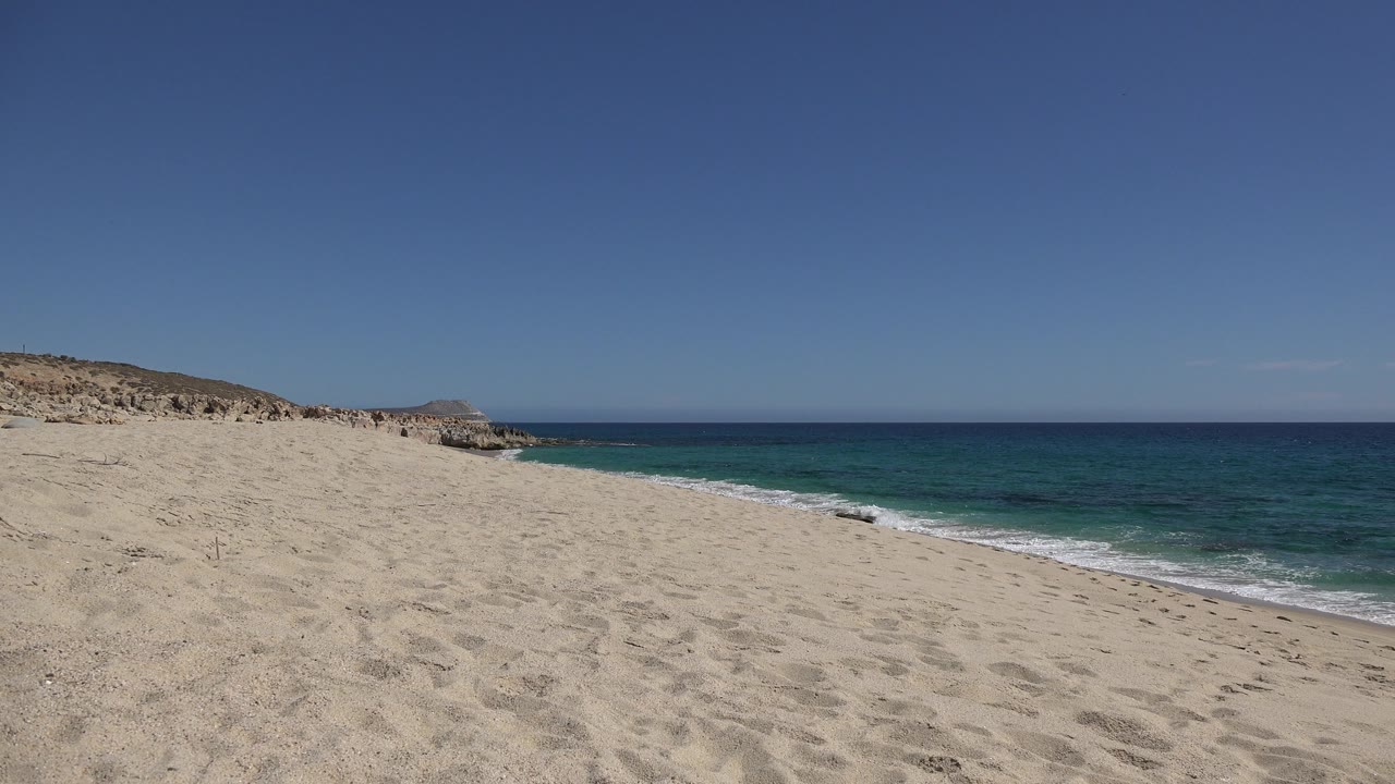 Early frame of the Los Cabos beach view with pale sand across the foreground, turquoise nearshore water at right, darker ocean beyond, and no visible people.