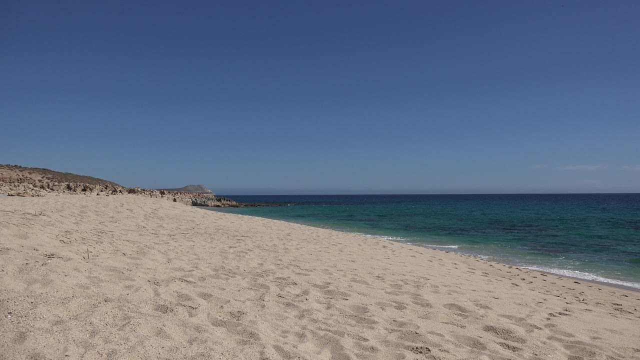 Opening frame of a Los Cabos sandy beach with footprints in the foreground, blue-green ocean at right, rocky coast at left, and no visible people.