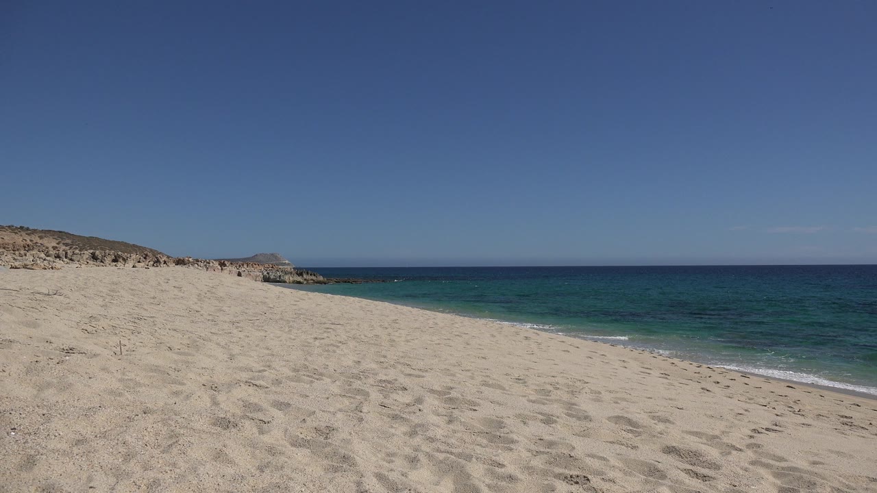 Los Cabos shoreline one second in, showing pale sand across the foreground, a low wave line on the right, rocky coast to the left, and no visible people.