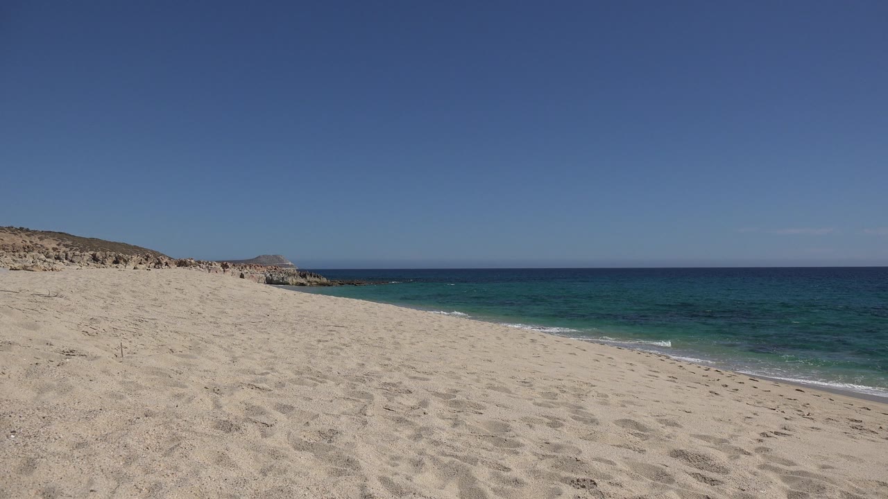 Empty beach with turquoise water meeting sand and low rocky land in the distance.