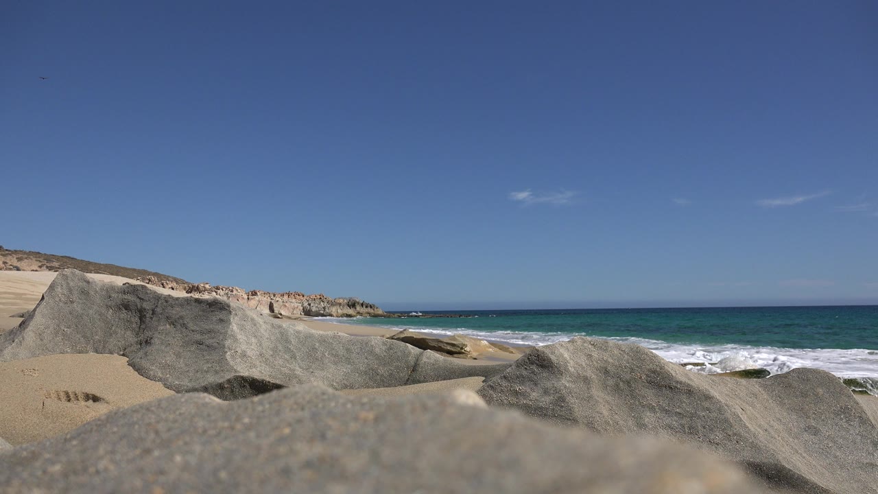 Opening frame of a Los Cabos shoreline with gray foreground rocks, pale sand at left, blue-green surf behind the rocks, distant tan bluffs, and no visible people.