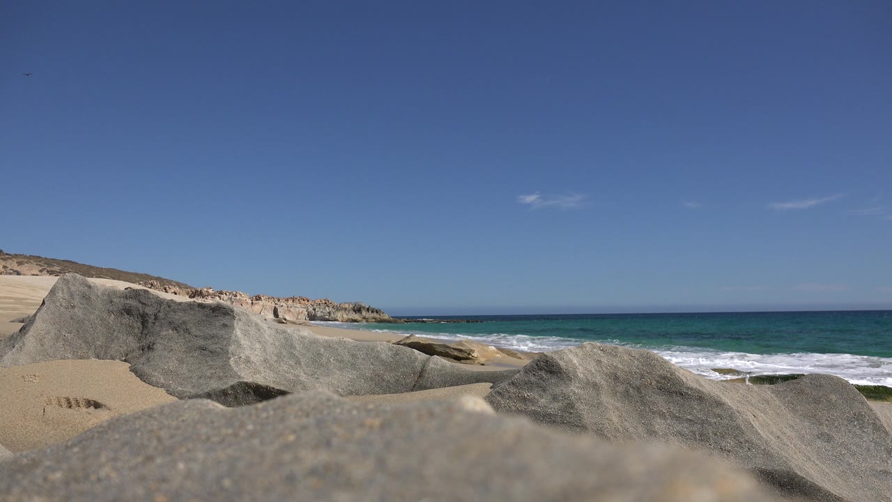 Ocean surf behind gray rocks with sandy shoreline and distant coastal bluffs.