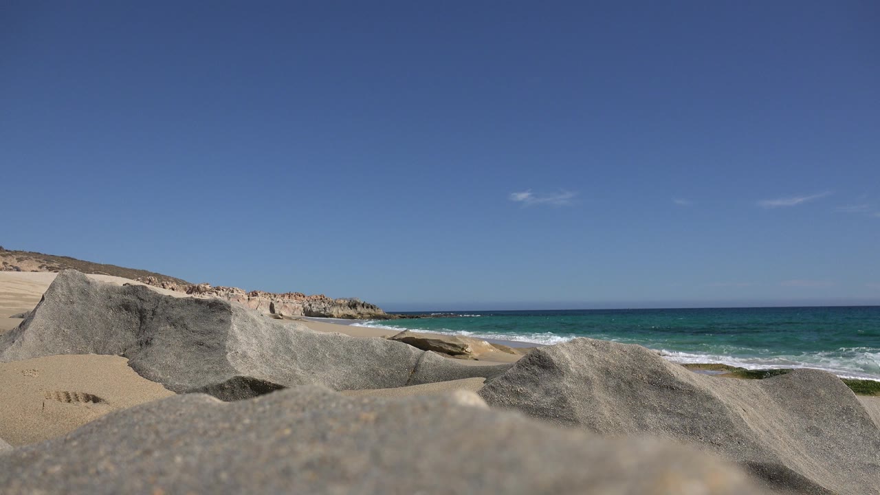 Opening frame of a Los Cabos beach with gray rocks close to the camera, tan sand at left, green-blue surf at right, distant low cliffs, and no visible people.