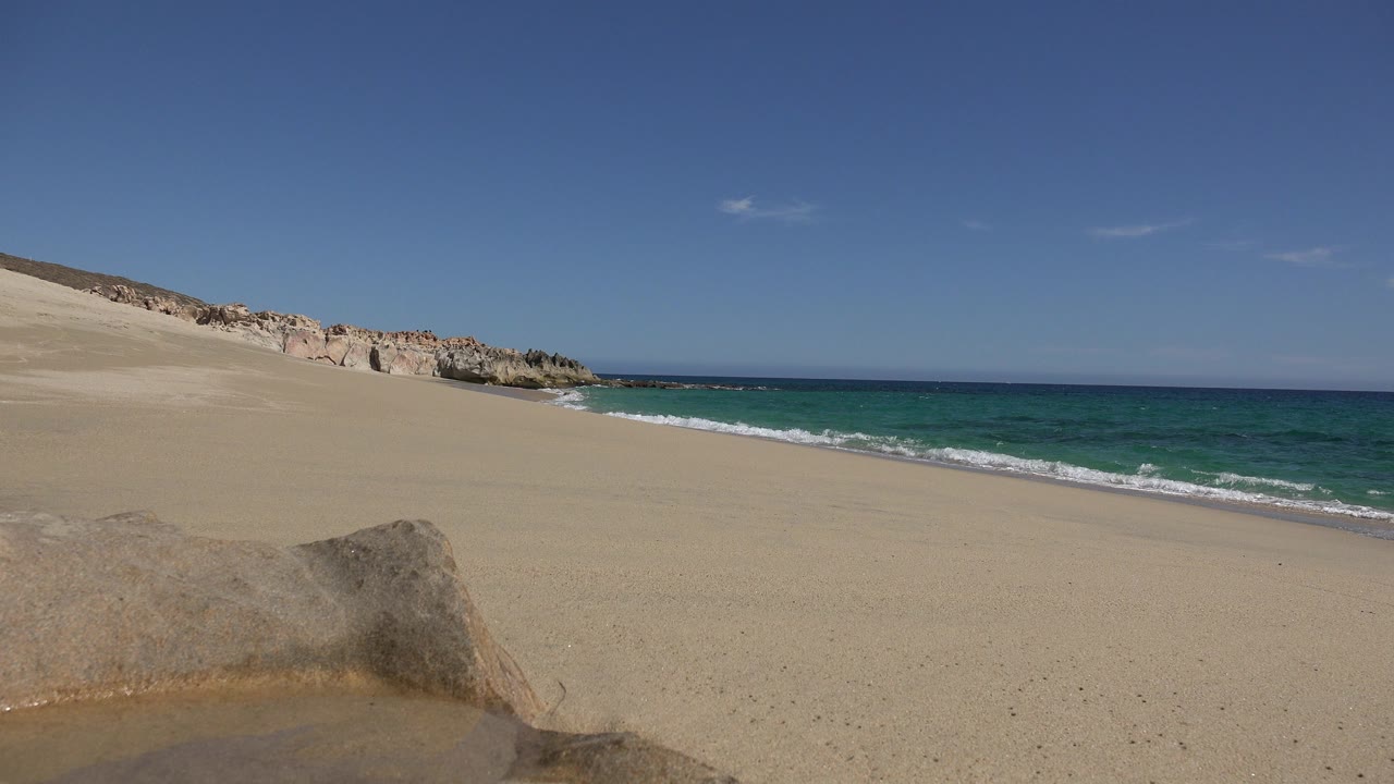 Opening view of a Los Cabos, Mexico beach with a large rock in the foreground, tan sand, blue-green water to the right, rocky shoreline at left, and no visible people.